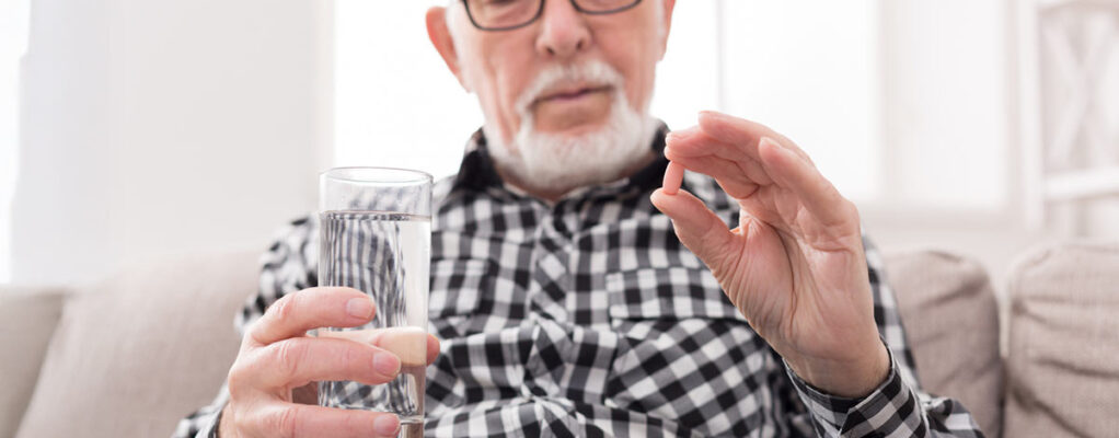 An older man holds a glass of water in one hand and a small pill in the other