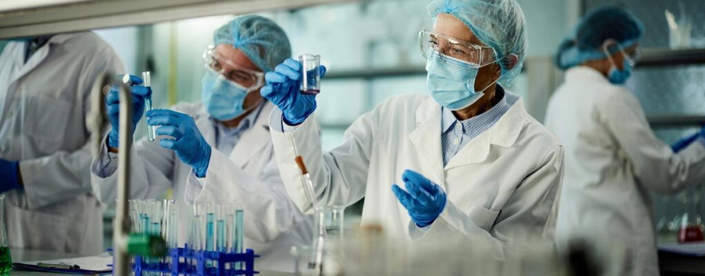 Researchers in protective lab gear, including masks, gloves, gowns, hairnets, and goggles, work in a laboratory setting.