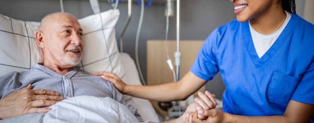 Nurse in scrubs smiling and holding the hand of an older male patient lying in a hospital bed during treatment