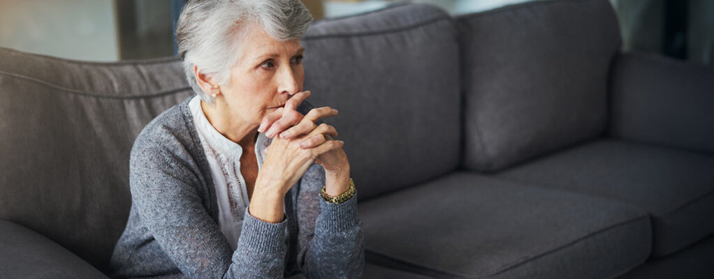 Elderly woman sitting on a couch, deep in thought, with her hands clasped in front of her face