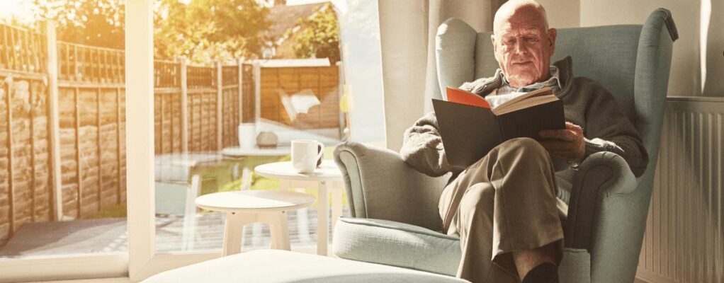 An older man sits in an armchair reading a book.